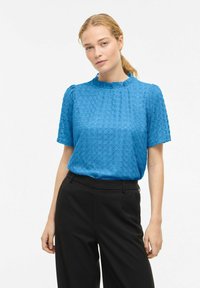 Blouse bleue texturée à manches courtes, col à volants et motif géométrique, associée à un pantalon noir. Finition en tissu lisse.