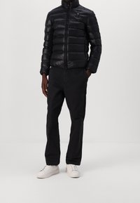 Een persoon in een zwarte pufferjack, zwarte broek en witte sneakers die tegen een effen lichte achtergrond staat.