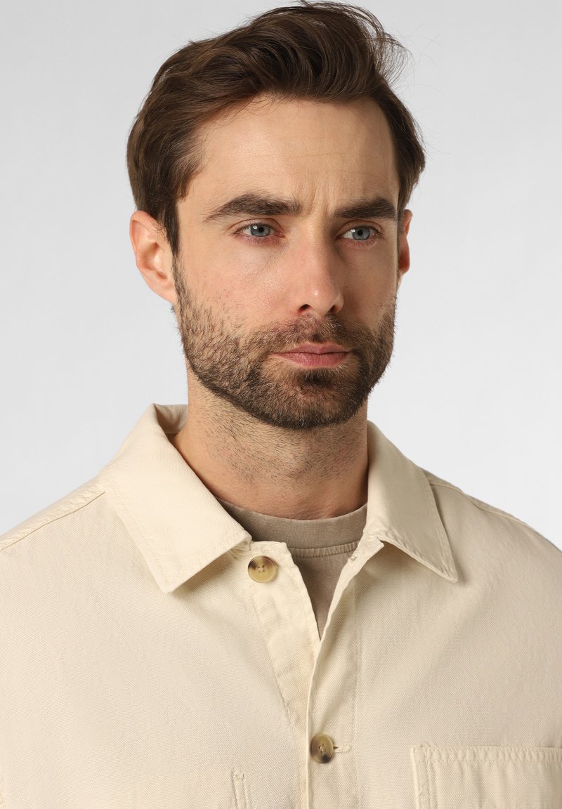 Chaqueta tipo camisa ligera de color beige con cuello, cierre de botones y bolsillo frontal, llevada sobre una blusa de un tono claro, con una tela texturizada.