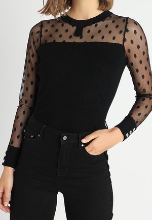 Femme portant un haut noir ajusté avec des manches longues transparentes à pois et un pantalon noir taille haute, main dans la poche.