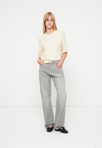 Pull léger couleur crème avec broderie florale, associé à un jean gris clair à jambes larges. Le modèle porte des ballerines noires, les mains dans les poches.