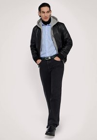 Černá kožená bomber bunda s šedou kapucí, světle modrá proužkovaná košile, tmavé džíny a černé boty. Zelený pásek přidává kontrastní detail.