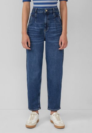 Osoba ubrana w niebieskie dżinsy z wysokim stanem i zwężanymi nogawkami, pasiastą niebieską koszulę oraz białe trampki, stojąca na szarym tle.