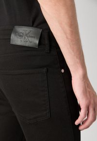 Los pantalones vaqueros negros de denim presentan una etiqueta de cuero en la cintura con "Calvin Klein Jeans" grabado. Incluyen bolsillos traseros estándar y detalles de costura.