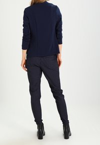 Granatowa kurtka z długim rękawem o gładkiej fakturze i detalem z tylnym szwem, zestawiona z granatowymi spodniami w prążki i czarnymi botkami za kostkę.