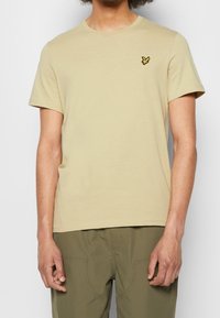 Jasnobeżowa bawełniana koszulka z okrągłym dekoltem, krótkimi rękawami oraz małym żółtym haftowanym logo na piersi. Miękka faktura, luźny krój.