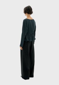 Top ribbed verde scuro con dettaglio raccolto sul retro e maniche lunghe, abbinato a pantaloni neri a gamba larga con vita alta. Texture liscia.