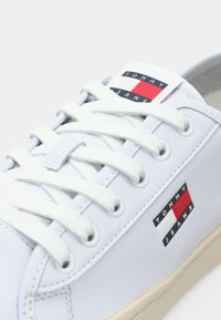 Valkoiset tennarit, jotka on valmistettu sileästä materiaalista, litteillä valkoisilla nauhoilla. Sivulla on punainen, valkoinen ja laivastonsininen logotagi sekä beige pohja.