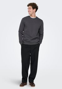 Grå sweatshirt med rund hals, kombineret med sorte løstsiddende bukser og brune slip-on sko, mod en ensfarvet lys baggrund.