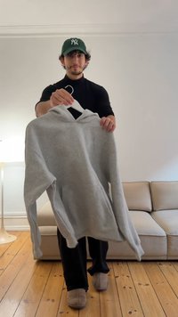 Grå hoodie med framficka, dragsko i huvan och ribbade muddar, upphängd på en vit galge mot en minimalistisk inomhusbakgrund.