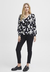 Blusa nera con motivi astratti bianchi e lilla chiaro, maniche lunghe a sbuffo, scollo a V, indossata con pantaloni aderenti neri e mocassini neri.