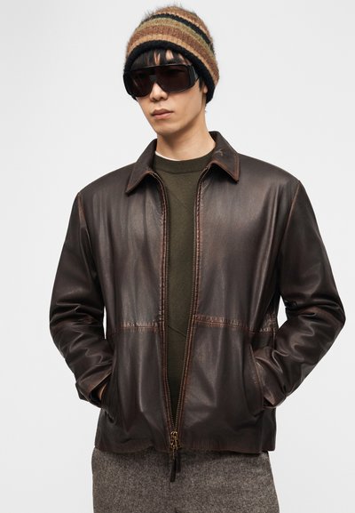 Άνδρας με σκούρο καφέ δερμάτινο μπουφάν, ελιά πράσινο πουλόβερ, μάλλινο καπέλο με ρίγες, μεγάλα μαύρα γυαλιά ηλίου και γκρι παντελόνι με τα χέρια στις τσέπες.