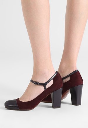 Woman’s legs wearing burgundy suede Mary Jane heels with black leather toe caps, ankle straps, and block heels on a white background.