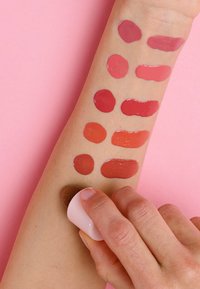 Muestras de diversos tonos de rojo y rosa para labios en un brazo de piel clara, mostradas sobre un fondo rosa, con una esponja para difuminar en uso.