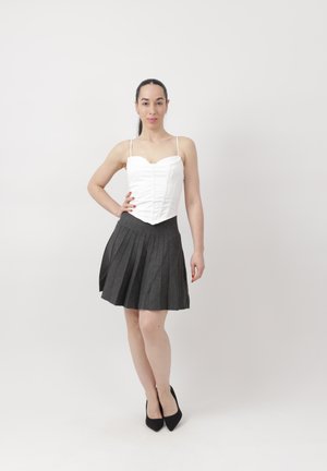 Vrouw staand met één hand op haar heup, gekleed in een wit corsettop, een donkere geplooide rok en zwarte hakken, tegen een effen witte achtergrond.