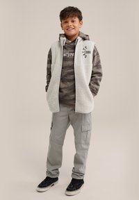 Pantalons cargo gris, un hoodie camouflage et un gilet gris clair avec des détails en polaire. Des baskets noires complètent l'ensemble.