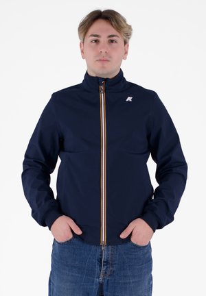 Giacca blu navy con colletto alto, chiusura frontale con zip caratterizzata da un accento arancione e polsini elasticizzati. Indossata con jeans azzurri.
