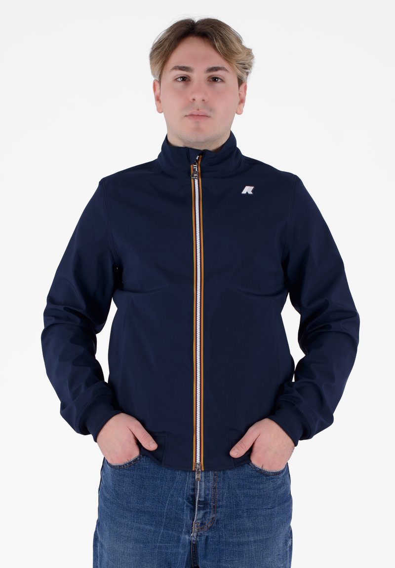 Giacca blu navy con colletto alto, chiusura frontale con zip caratterizzata da un accento arancione e polsini elasticizzati. Indossata con jeans azzurri.
