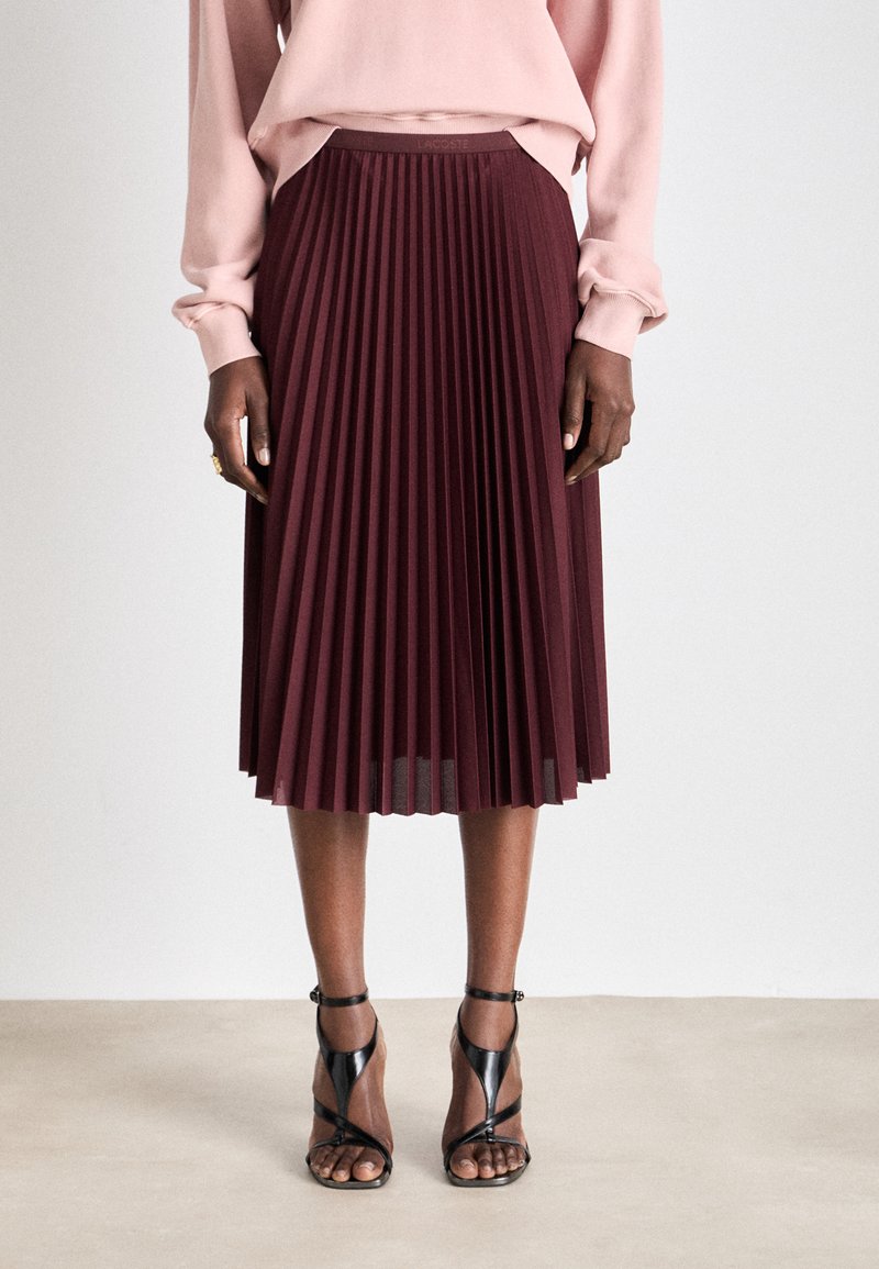 Falda plisada burdeos con una cinturilla suave, combinada con un suéter rosa claro y sandalias de tacón alto negras con tiras, de pie sobre un suelo claro.