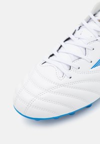 Zapatilla de fútbol blanca con parte superior de cuero texturizado, que presenta acentos azules en el costado y una suela de color azul claro con tapones moldeados.