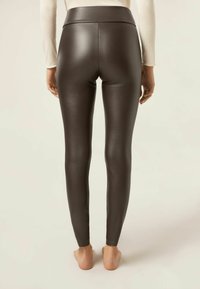 Bruna leggings i konstläder med hög midja, slät textur och tight passform, sedda bakifrån med bara fötter synliga.