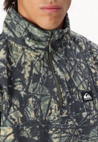 Fleece-pulloveissa on maastokuvio, jossa on korkea kaulus ja puoli vetoketju, harmaan, vihreän ja beigeen sävyissä, varustettuna bränditunnuksella.