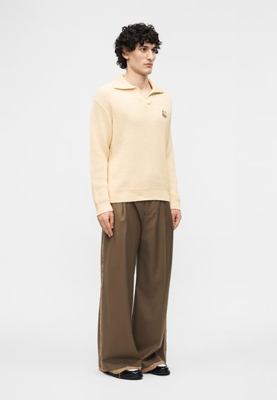 Homme aux cheveux bouclés portant un pull en maille crème et un pantalon marron ample, debout devant un fond clair uni.