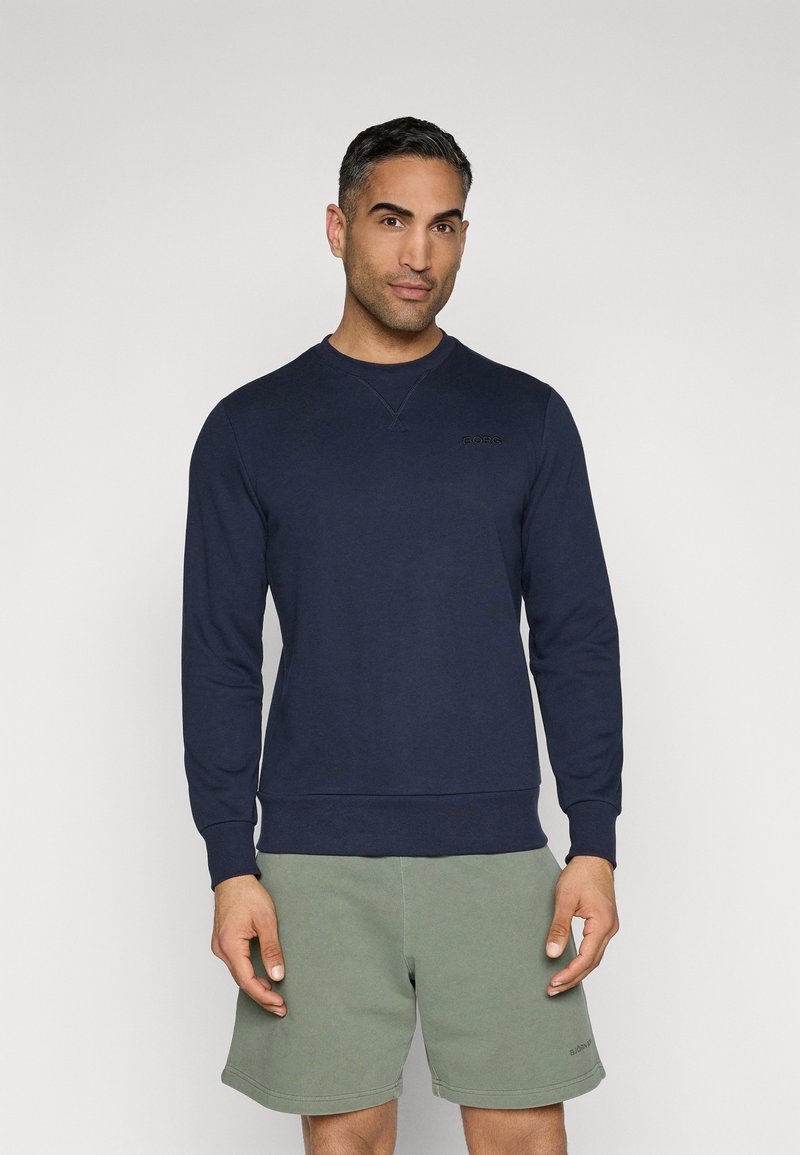 Felpa blu navy con scollo tondo e maniche lunghe, con un discreto dettaglio del logo. Abbinata a pantaloni corti verde chiaro.