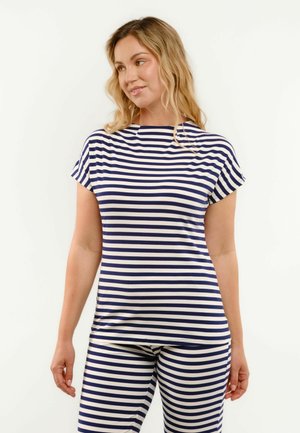 Kobieta o falujących blond włosach, ubrana w granatowo-biały top w poziome paski z krótkim rękawem oraz dopasowane spodnie, stojąca na białym tle.