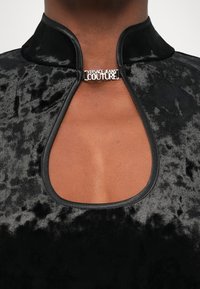 Crni baršunasti top s visokim ovratnikom i izrezom u obliku ključa, s srebrnim logotipom koji kaže "Versace Jeans Couture."