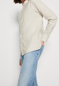 Beige Langarmshirt mit Rundkragen und Knopfleiste, kombiniert mit hellblauen Jeans. Besitzt eine glatte Textur und eine schmale Passform.