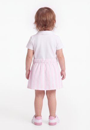 Tout-petit debout de dos, portant un t-shirt blanc à manches courtes, une jupe rayée rose et blanche, et des baskets blanches avec des touches roses.