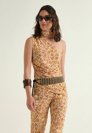 Femme portant un top asymétrique à motifs et un pantalon assorti, des lunettes de soleil foncées, des boucles d'oreilles à pompons, une large ceinture rayée et un bracelet manchette en bois.