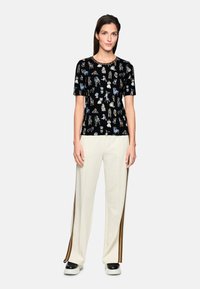 Black t-shirt with colorful patterns of figures, short sleeves, round neckline. Paired with cream-colored trousers featuring side stripes.