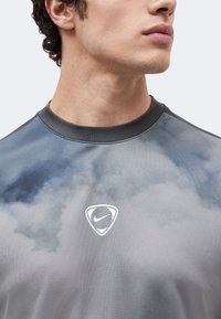 Νέος άνδρας που φοράει γκρι και μπλε φανέλα Nike tie-dye με μαύρο γιακά, κοιτάζοντας δεξιά μπροστά σε ένα απλό φωτεινό φόντο.