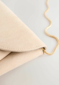 Beige Wildleder-Clutch mit einer umgeklappten Klappe und einem dünnen goldenen Kettenriemen auf einer hellen Oberfläche.