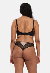 Femme aux cheveux tressés portant un soutien-gorge en dentelle noire et un string en dentelle transparente assorti, debout contre un fond clair uni, vue de dos.