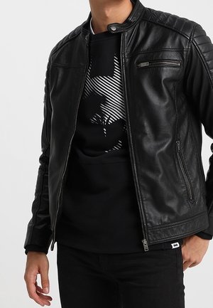 Homme portant une veste en cuir noire par-dessus un sweat-shirt graphique noir avec un logo Batman composé de lignes horizontales.