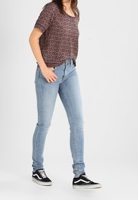 Frau, die ein gemustertes kurzärmliges Hemd, hellblaue Skinny Jeans und schwarze Low-Top-Sneaker trägt, steht vor einem einfarbigen Hintergrund.