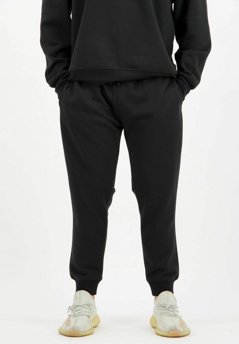 Schwarze Sweatpants mit elastischem Bund, Bündchen an den Knöcheln und Seitentaschen, kombiniert mit hellen Sneakers. Weiche Stoffstruktur.