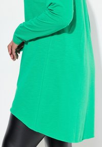 Grüne langärmlige Tunika aus weichem Stoff mit glatter Textur, asymmetrischem Saum, kombiniert mit schwarzen Leggings.