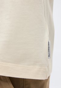 Μπεζ ύφασμα με ρέλι και μια μικρή λευκή ετικέτα που γράφει "Marc O'Polo Denim" ραμμένη στην πλαϊνή ραφή πάνω από καφέ παντελόνια.