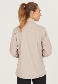Lys beige jakke med høj krave, lavet af glat stof. Har raglanærmer og en buet kant for en afslappet pasform. Ingen synlig hardware.