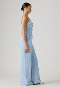 Γυναίκα με σκούρα μαλλιά σε κοτσίδα, που φοράει ανοιχτό μπλε τζιν ολόσωμη φόρμα με φαρδιά πόδια και λευκά αθλητικά παπούτσια, στέκεται πλαγίως σε απλό φόντο.