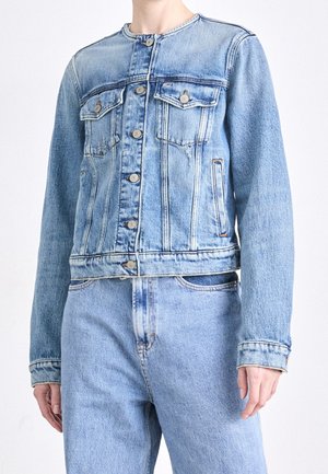 Person, die eine hellblaue, geknöpfte Denimjacke mit Brusttaschen und passende hoch taillierte Jeans vor einem einfarbigen Hintergrund trägt.