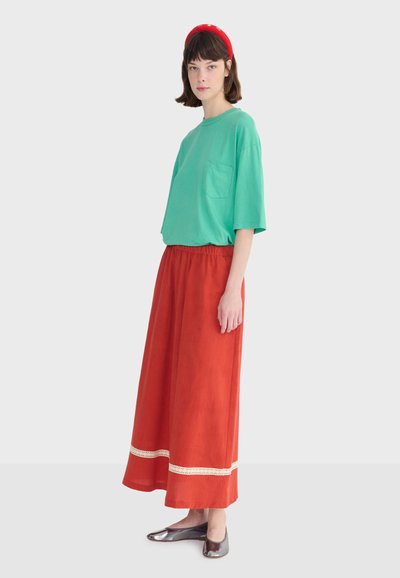 Femme portant un t-shirt vert oversize, une jupe maxi rouge avec bordure blanche, des ballerines métalliques et un bandeau rouge, debout de profil sur un fond blanc.