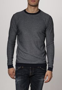 Grijze langmouwen sweater met geribbelde marineblauwe halslijn en manchetten. Beschikt over een textuurpatroon van gemêleerde stof. Gedragen met donkere spijkerbroeken.
