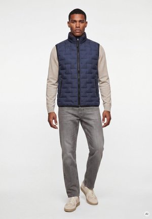 Mann trägt eine marineblaue gesteppte Weste über einem beigen Langarmshirt, graue Jeans und beige Sneakers und steht vor weißem Hintergrund.
