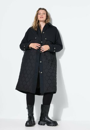 Donna che indossa un lungo cappotto trapuntato nero, leggins neri e stivali neri chunky, in piedi su uno sfondo bianco.