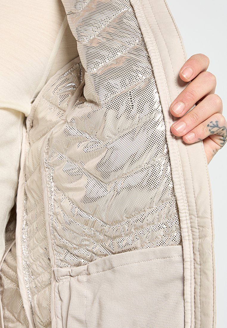 Licht beige jack met een gequilte zilveren binnenkant, met gaasdetails en een zak, die een gestructureerd uiterlijk vertoont. Hand gedeeltelijk zichtbaar.